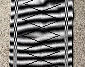 Puede incluir: Alfombra rectangular en blanco y negro con un patrón geométrico de diamantes. La alfombra presenta rayas horizontales y un borde con un diseño detallado. La alfombra está hecha de un material texturizado.