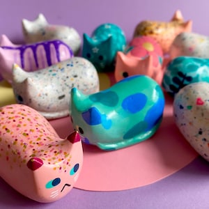 May include: A collection of small, smooth, cat-shaped figurines in various colours and patterns. The figures feature designs including multi-coloured speckles, solid bright blue spots, purple drips, and floral motifs, all with painted facial features.