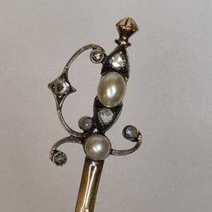 Op de afbeelding: Een sierlijke, antieke haarspeld met een goudkleurige schacht en een decoratieve bovenkant. De bovenkant heeft twee parels, verschillende heldere edelstenen en een goudkleurige eindknop. Het ontwerp is symmetrisch en elegant, met een vintage esthetiek.