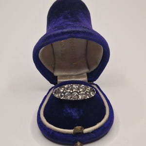 May include: An oval-shaped ring with multiple clear stones set in a silver-colored band, displayed inside a vintage blue velvet ring box. The box is open, revealing the ring against a cream-colored interior.