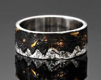 May include: A silver band ring with a black and gold textured design. The design features a mountain range with gold accents.