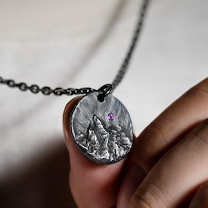 May include: A dark, oxidized silver pendant on a matching chain. The circular pendant features a textured, relief-style mountain landscape design with a single, small, round, vibrant purple gemstone embedded near the top edge of the metal surface.