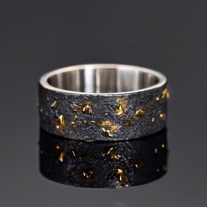 May include: A wide, textured ring with a dark gray exterior and gold-colored accents. The interior of the ring is a bright silver color. The ring is displayed on a reflective black surface.