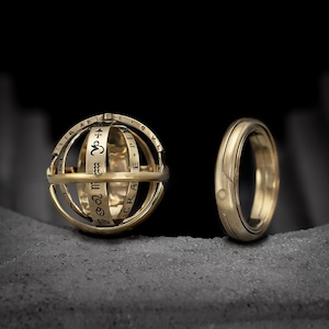 Op de afbeelding: Twee goudkleurige ringen. Eén is een bol met dierenriemtekens en banden. De andere is een effen band met een klein cirkelvormig ontwerp. Beide ringen liggen op een grijze ondergrond tegen een donkere achtergrond.