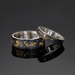Könnte beinhalten: Zwei Silberringe mit einem strukturierten schwarzen Band und goldenen Akzenten. Die Ringe werden auf einer reflektierenden Oberfläche präsentiert, die ihr detailliertes Design hervorhebt. Diese Ringe sind wahrscheinlich für eine Hochzeit oder einen Verlobungsring.