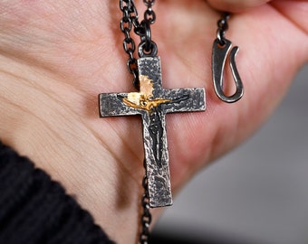 May include: A dark, oxidised silver cross pendant featuring a textured, hammered surface and a central, raised gold-toned figure. The cross hangs from a matching dark silver chain with a hook-style clasp, showcasing a rugged, industrial aesthetic.