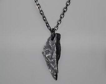 May include: A dark grey pendant necklace with a textured, jagged pendant. The pendant hangs from a matching chain, showcasing a handcrafted, rustic aesthetic. The necklace design highlights the pendant's unique texture and irregular shape.