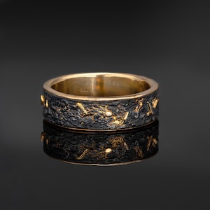 Op de afbeelding: Een goud-zwarte ring met een getextureerde band. De ring heeft een donker, ruw exterieur met gouden accenten en een glad, gepolijst interieur. De ring wordt gepresenteerd op een reflecterend oppervlak.
