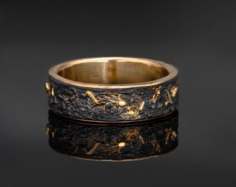 May include: A gold and black ring with a textured band. The ring has a dark, rough exterior with gold accents and a smooth, polished interior. The ring is displayed on a reflective surface.