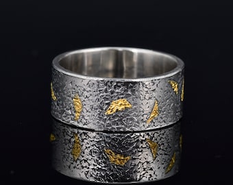May include: A silver ring with a textured surface and gold-colored accents. The ring has a wide band and a pattern of irregular gold shapes. The ring is displayed on a reflective black surface.