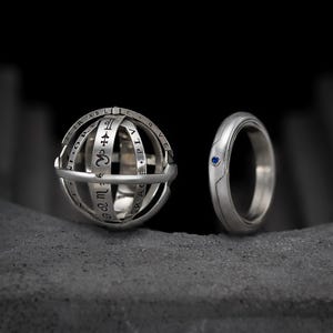 Peut inclure: Deux bagues en argent sur une surface grise. L'une est une sphère avec des symboles et du texte gravés. L'autre est une simple bague avec une petite pierre bleue. Les bagues sont posées sur un fond sombre.