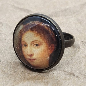 Puede incluir: Un anillo con un camafeo redondo de vidrio que presenta un retrato de una mujer con cabello castaño. El camafeo está engastado en un anillo de metal de color bronce.