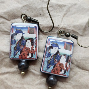 Op de afbeelding: Een paar rechthoekige oorbellen met een Japanse kunstdruk. De print toont twee figuren in traditionele kleding. Elke oorbel heeft een donkergrijze kraal en een kleine gouden kraal. De oorbellen hebben antiek messingkleurige oorhaken.