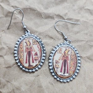 May include: A pair of silver earrings with oval-shaped pendants featuring an image of an Egyptian goddess with hieroglyphics. The pendants are surrounded by a row of silver beads.