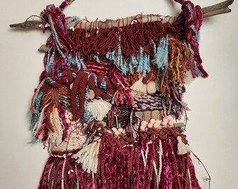 May include: A handmade wall hanging featuring a variety of textures and colors. The piece incorporates burgundy, teal, and cream-colored yarns, along with a wooden branch and a twisted rope for hanging. The design includes tassels and decorative elements.