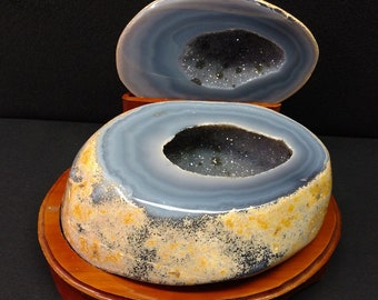May include: A geode with a large, open cavity lined with sparkling crystals. The geode is a light blue color with a brown outer layer and is displayed on a wooden base.