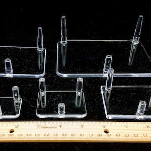 May include: Five clear acrylic display stands of varying sizes, each with three prongs, on a wooden ruler. The ruler is marked in inches.