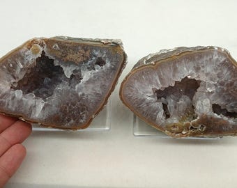 May include: Two halves of a geode, showcasing a natural crystal formation. The outer edges are a mix of brown and tan, while the interior reveals a sparkling, white crystal structure. The geodes are displayed on clear stands.
