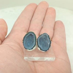 Op de afbeelding: Twee blauwe geode oorbellen met een donkerblauwe rand en witte basis worden gepresenteerd op een heldere, rechthoekige standaard. De oorbellen hebben een sprankelend, kristalachtig interieur. De oorbellen worden in een hand gehouden.