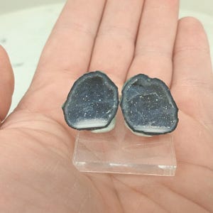 Op de afbeelding: Twee donkerblauwe geode schijfjes met witte kristallen interieurs worden gepresenteerd op een heldere acryl standaard. De geodes hebben een donkere rand en een witte basis.