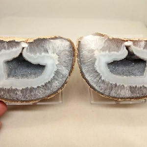 May include: Two halves of a geode, showcasing a natural stone formation. The outer edges are a light tan, transitioning to a gray and white interior with a crystalline center. The geode halves are displayed on clear stands.