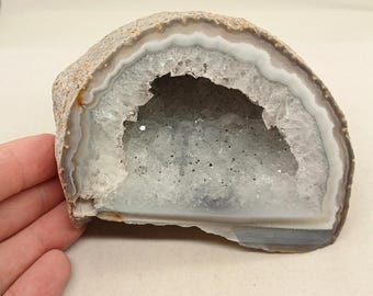 May include: A semi-circular agate geode featuring a rough, tan-colored outer crust and a hollow center lined with clear, sparkling quartz crystals. The interior cavity is surrounded by distinct, concentric bands of white and light gray mineral layers.