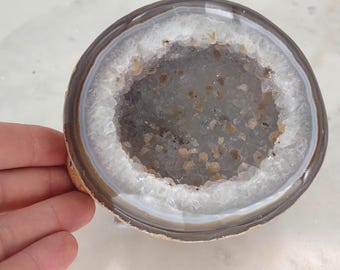 May include: A round, white agate geode with a brown outer edge. The geode has a hollow center with a rough, gray surface.