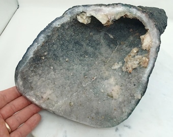 May include: A large, open geode with a dark gray exterior and a light gray interior filled with small, crystal-like formations. The geode's cavity reveals white and tan crystalline structures. The overall shape is irregular and organic.