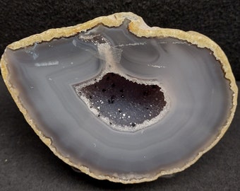 May include: A gray agate geode with a large, open cavity lined with sparkling crystals. The geode has a smooth, polished exterior with a light brown rim.