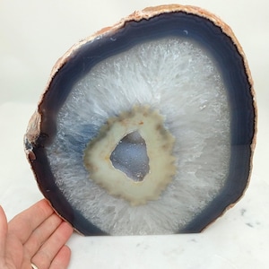 May include: A polished agate geode slice with a natural, rough edge. The interior features concentric bands of white, blue, and grey, with a central cavity. The geode is a decorative item, perfect for display.