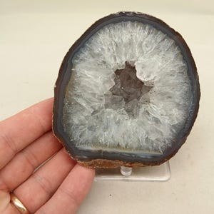 以下が含まれることがあります： 透明でギザギザした水晶の結晶で満たされた空洞の中心部を持つ、磨かれた瑪瑙のスライス。石の外縁は暗いアースブラウンで、輝く半透明の白い内部を縁取る薄い灰色の帯へと変化しています。