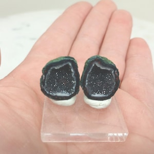 Op de afbeelding: Twee kleine, geode-achtige objecten met donkere interieurs en groene en rode randen worden getoond. Ze liggen op een heldere, rechthoekige basis, vastgehouden in een hand. De geodes hebben een witte basis.