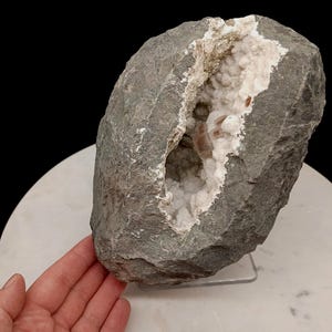 May include: A large, rough, dark gray geode rock featuring a central cavity filled with white crystalline structures and small, pale pinkish-brown mineral formations. The exterior of the stone has a textured, uneven surface with natural fissures.