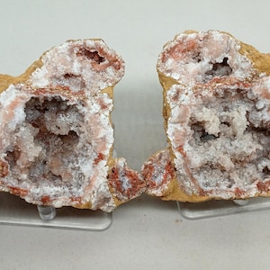 May include: Two halves of a geode, showcasing a natural stone formation. The exterior is a tan color, while the interior reveals a crystalline structure with shades of white, pink, and red. The geode is displayed on a clear stand.