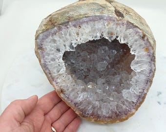 May include: A large, open geode with a rough, tan exterior and a cavity lined with sparkling, clear quartz crystals. The interior has a purple hue around the edges. The geode is partially held by a hand.