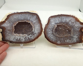 May include: Two halves of a geode, showcasing a natural formation. The outer edges are a warm brown, transitioning to a white and gray crystalline interior. The center reveals a darker, crystal-filled cavity. Each piece is displayed on a clear stand.