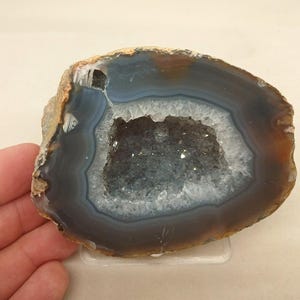 以下が含まれることがあります： 濃い灰色、青、茶色の同心円状の帯が特徴の磨かれた瑪瑙のスライス。中央の空洞になったジオードの空洞には、小さく輝く半透明の水晶の結晶が詰まっており、外縁は粗く自然な質感になっています。