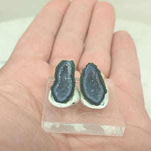 Op de afbeelding: Twee kleine, gepolijste geodehelften met een donkerblauwe binnenkant en een witgroene buitenrand. Ze worden gepresenteerd op een heldere, rechthoekige basis, die hun kristalstructuur benadrukt. De foto is gemaakt in natuurlijk licht.