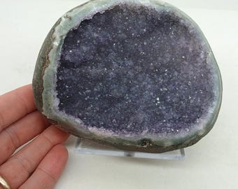 May include: A natural amethyst geode, showcasing a deep purple crystal interior. The outer edge is a mix of gray and green hues, with a polished appearance. The geode is displayed on a clear stand, highlighting its natural beauty.