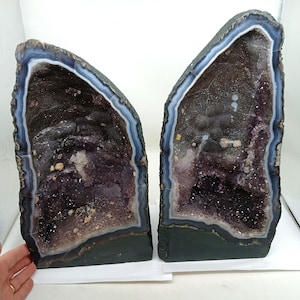 Op de afbeelding: Twee amethist geode boekensteunen. De geodes hebben een donkerpaars interieur met kristalformaties en een donkerblauwe en witte rand. De buitenste randen zijn donkergrijs van kleur. De boekensteunen worden op een wit oppervlak weergegeven.
