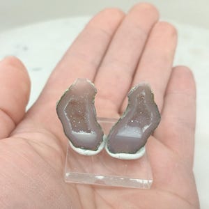 Op de afbeelding: Twee bijpassende, gepolijste geodehelften, met een lichtpaars interieur met sprankelende kristalformaties. De geodes zijn gemonteerd op een heldere, rechthoekige basis, met een groen-witte rand. De algehele esthetiek is natuurlijk en elegant.