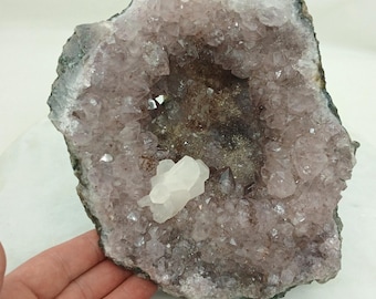 May include: A large amethyst geode with a hollow center, showcasing a cluster of purple crystals. A smaller, white quartz crystal sits inside the geode. The geode's exterior is a dark gray color.