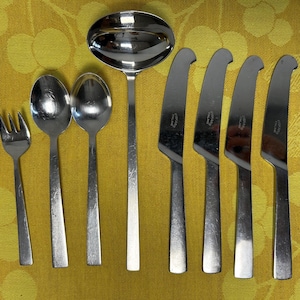 Op de afbeelding: Een set roestvrijstalen bestek, waaronder een vork, twee lepels, een pollepel en vier messen. Het bestek is gerangschikt op een gele tafelkleed met patroon. De messen hebben een gebogen mesontwerp.