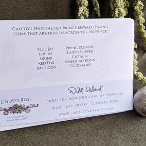 May include: A white card with blue text lists ten Prince Edward Island items, including blue jay, lupins, skunk, red fox, raccoon, piping plovers, lady's slipper, cattails, American robin, and chickadee. The card also features the text "Wild Island" and "Lindsey Ross, a non-traditional traditional artist".
