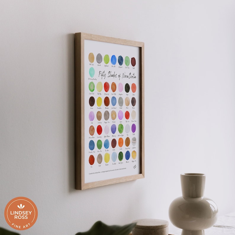 May include: A framed print titled "Fifty Shades of Nova Scotia" displaying a grid of 50 watercolour circles in various shades of blue, green, red, orange, yellow, brown, purple, and grey. Each circle is labelled with a name, such as "Sea Glass", "Fog", "Red Clay", and "Sand".