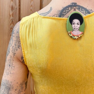 May include: A yellow sleeveless shirt with a green and brown oval pin featuring a portrait of a woman with an afro.