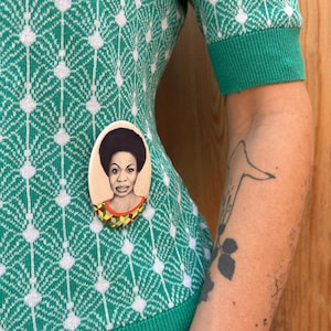 May include: A green and white patterned sweater with a round pin featuring a portrait of a woman with dark hair and a colorful patterned shirt.
