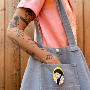 May include: A gray canvas tote bag with a yellow pocket. A round pin with a colorful portrait of a woman is attached to the pocket.