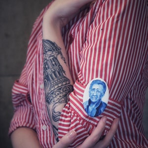 May include: A person wearing a red and white striped shirt with a black and white tattoo on their arm. They are holding a white oval pin with a blue portrait of a person wearing glasses.