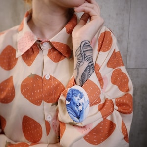 May include: A white button-down shirt with a strawberry print. The shirt has a blue and white oval brooch with a portrait of a woman.